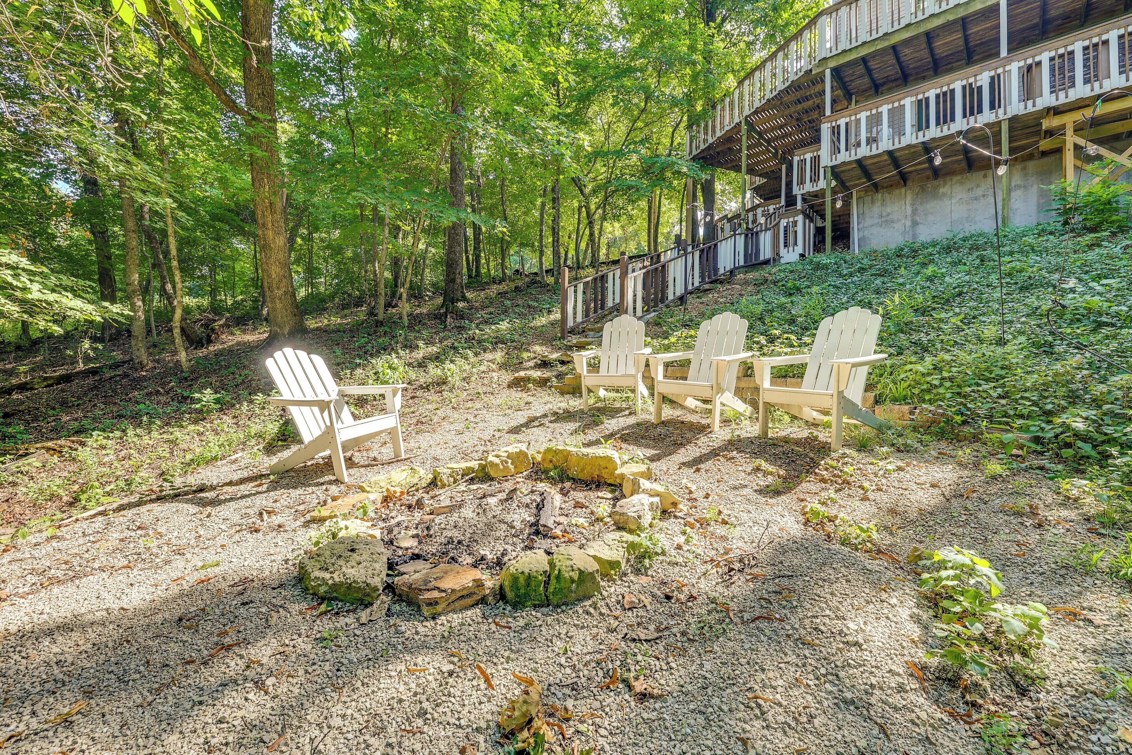 A forested area with a fire pit and Adirondack chairs.