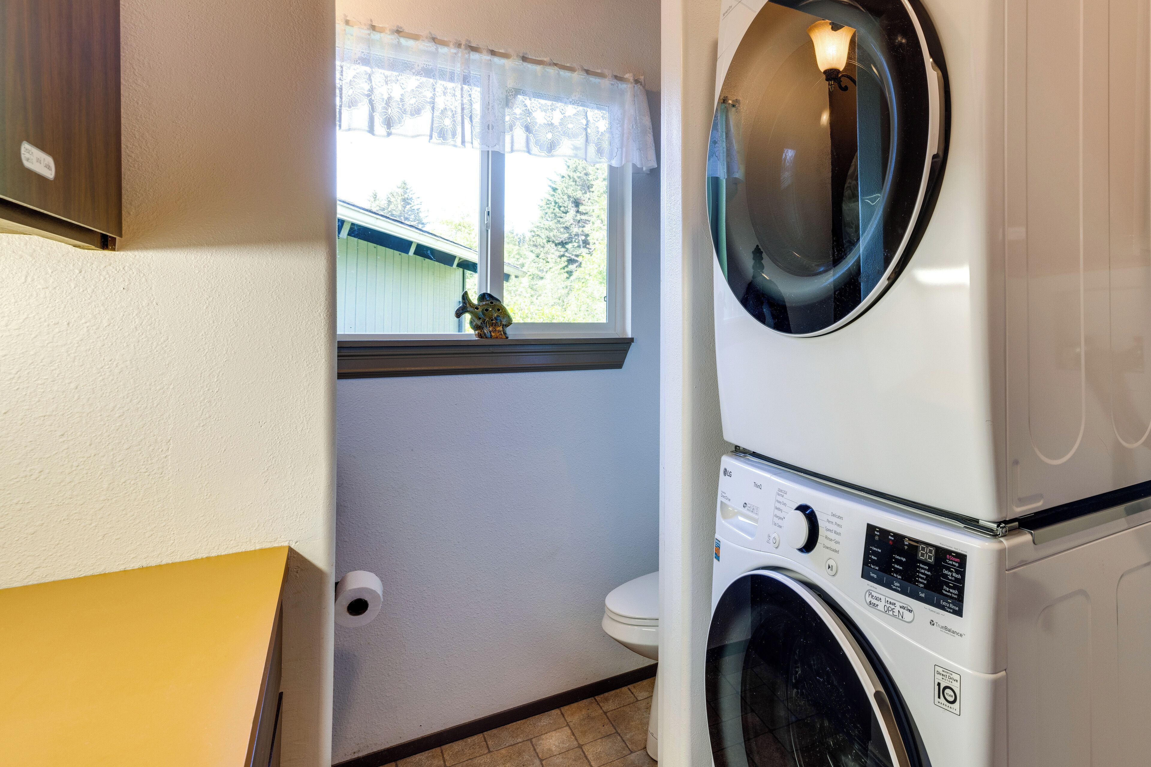 Une petite buanderie comprenant une machine à laver et un sèche-linge superposés, une fenêtre donnant sur un espace verdoyant et des toilettes.