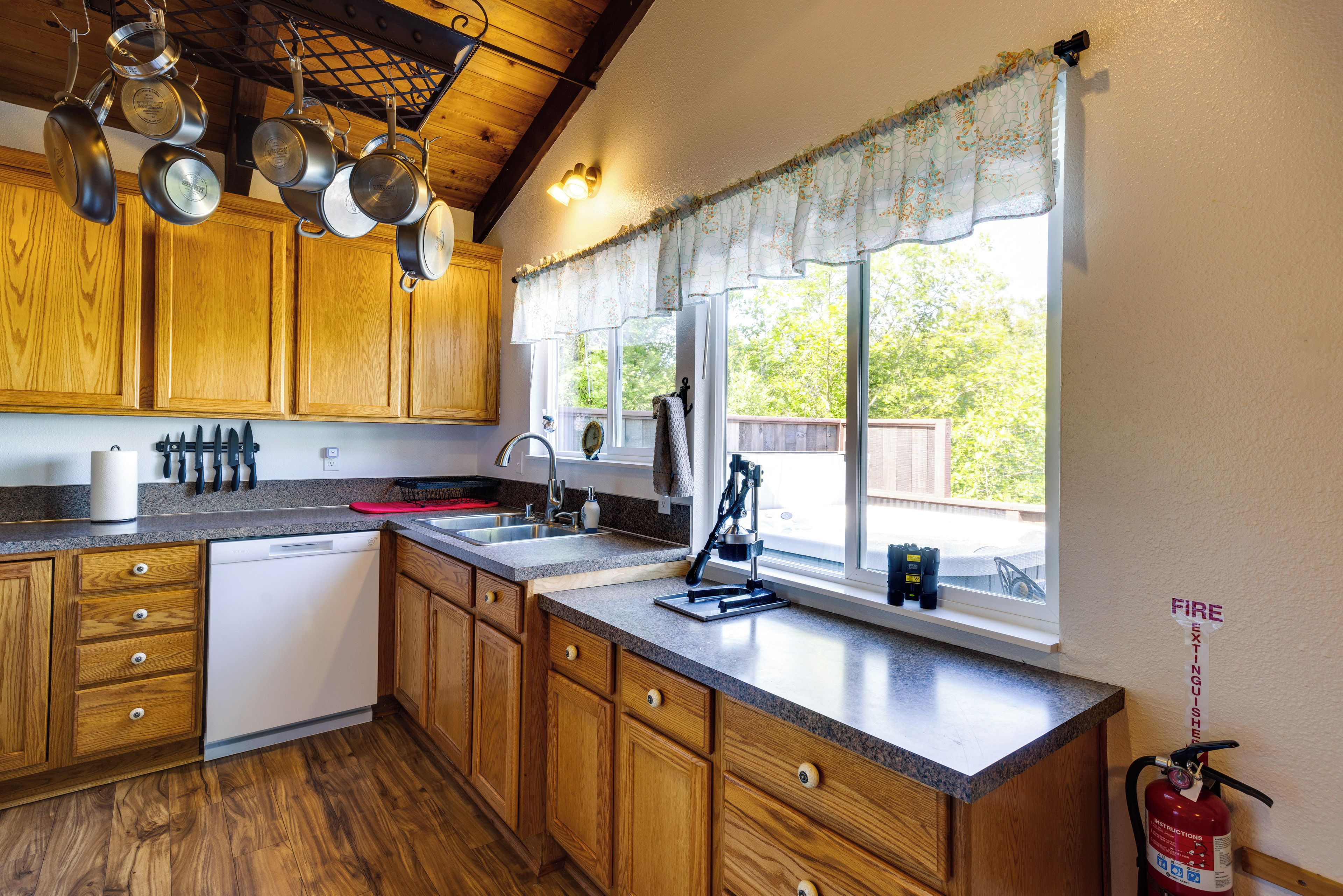 A kitchen with wooden cabinets, a dishwasher, a window with a view of greenery, and a fire extinguisher mounted on the wall.