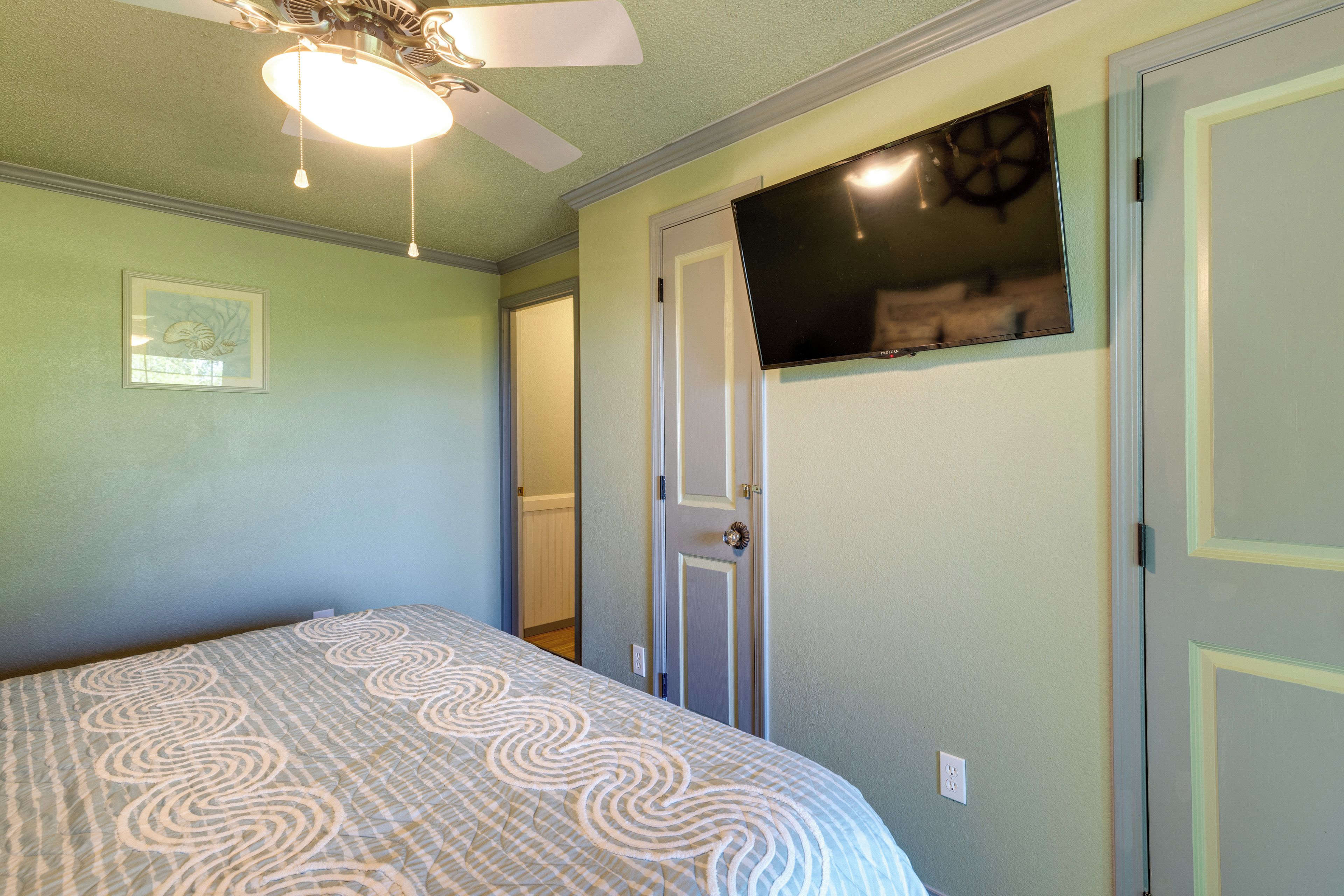 A bedroom with a bed, a ceiling fan, a TV mounted on the wall, and a door leading to another room.