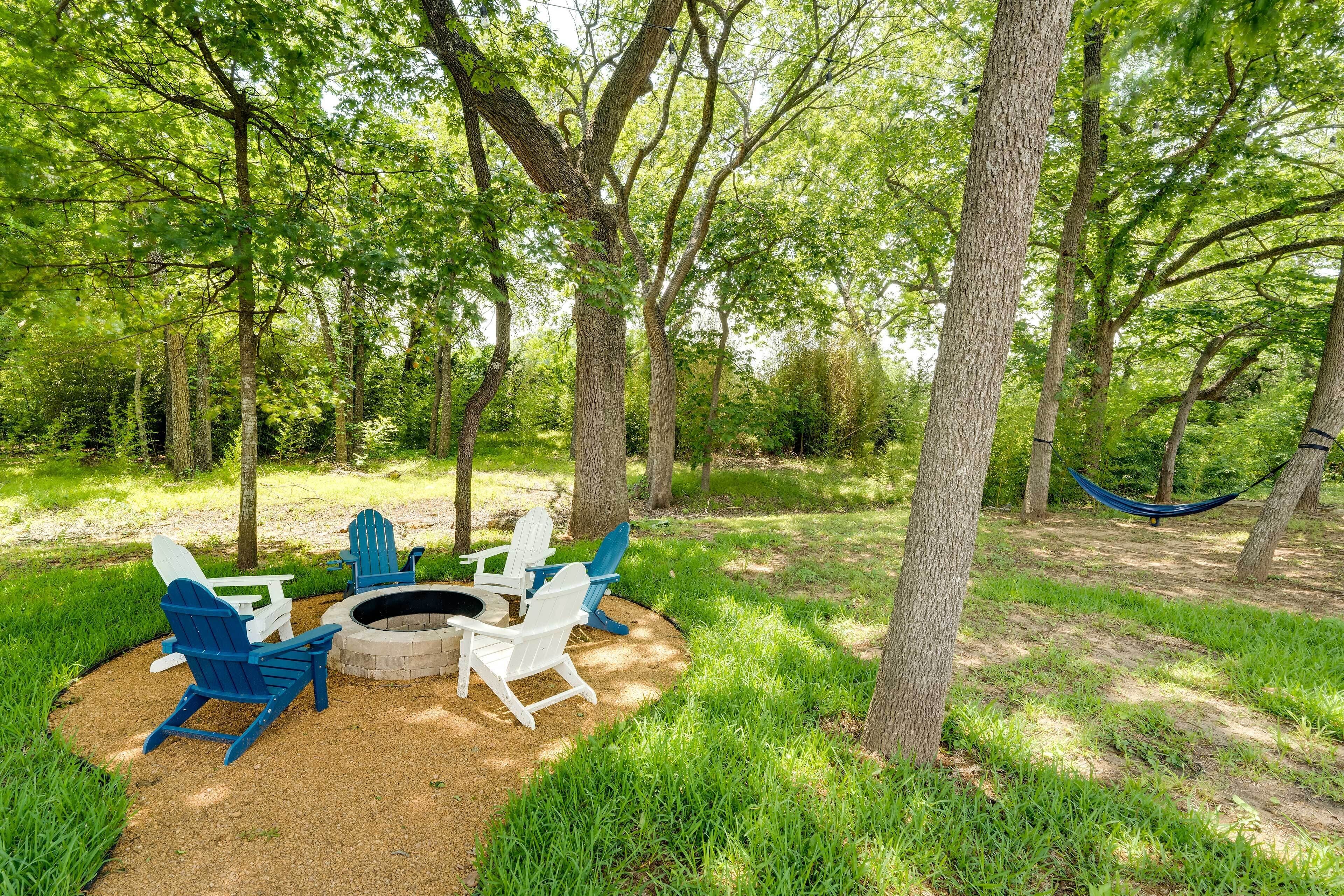 A forested area with a fire pit, white and blue Adirondack chairs, and a hammock.