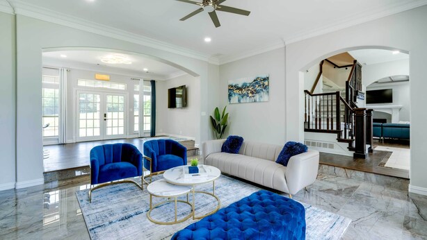 A spacious living room with a white sofa, blue armchairs, a blue ottoman, and a white coffee table. There is a staircase leading to another level, and a painting on the wall.