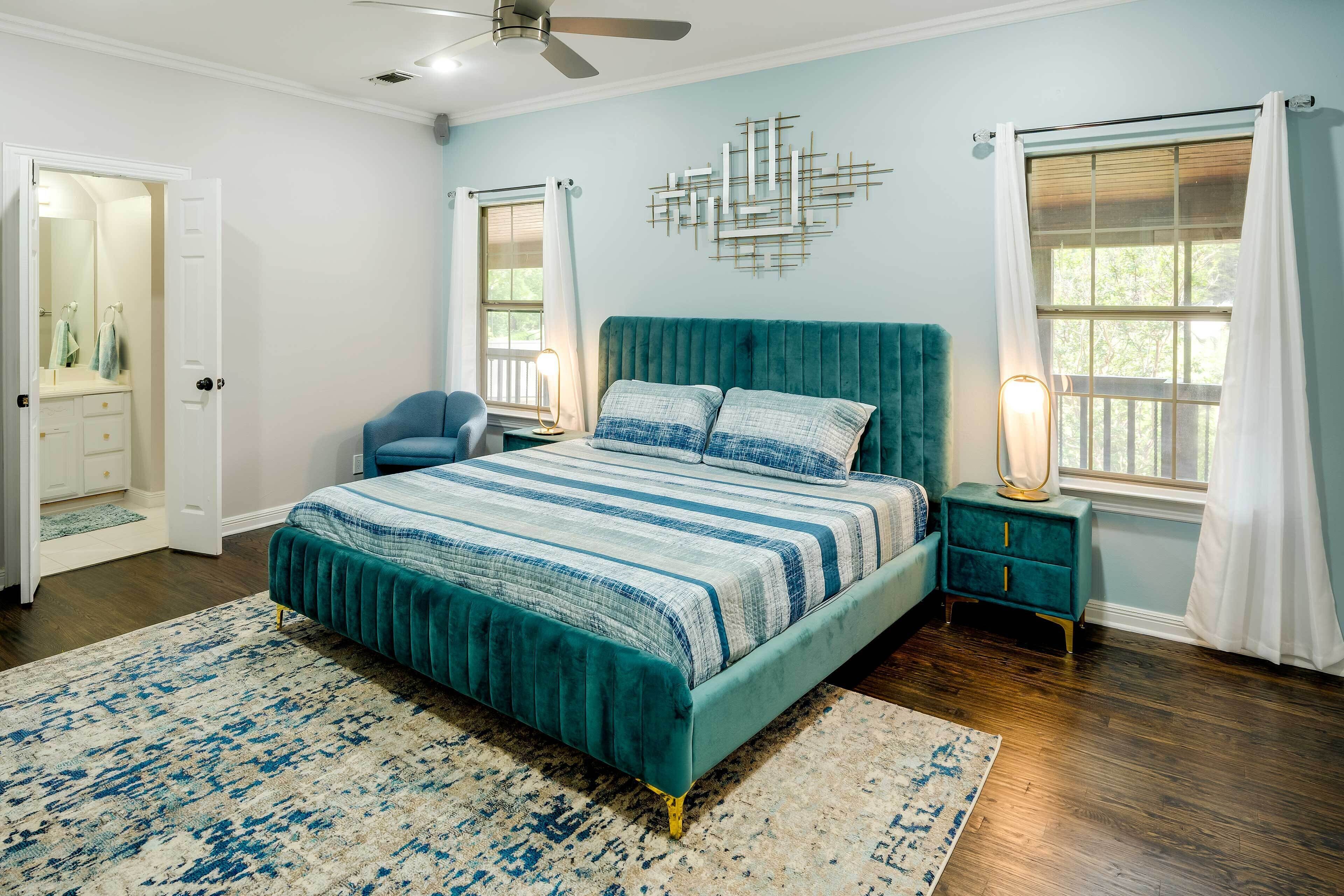 Ein Schlafzimmer mit einem turquoise-farbenen Bett, einer blau-weißen gestreiften Tagesdecke, einem weißen Teppich, einem Nachttisch mit Lampe und einem Fenster mit weißen Vorhängen.