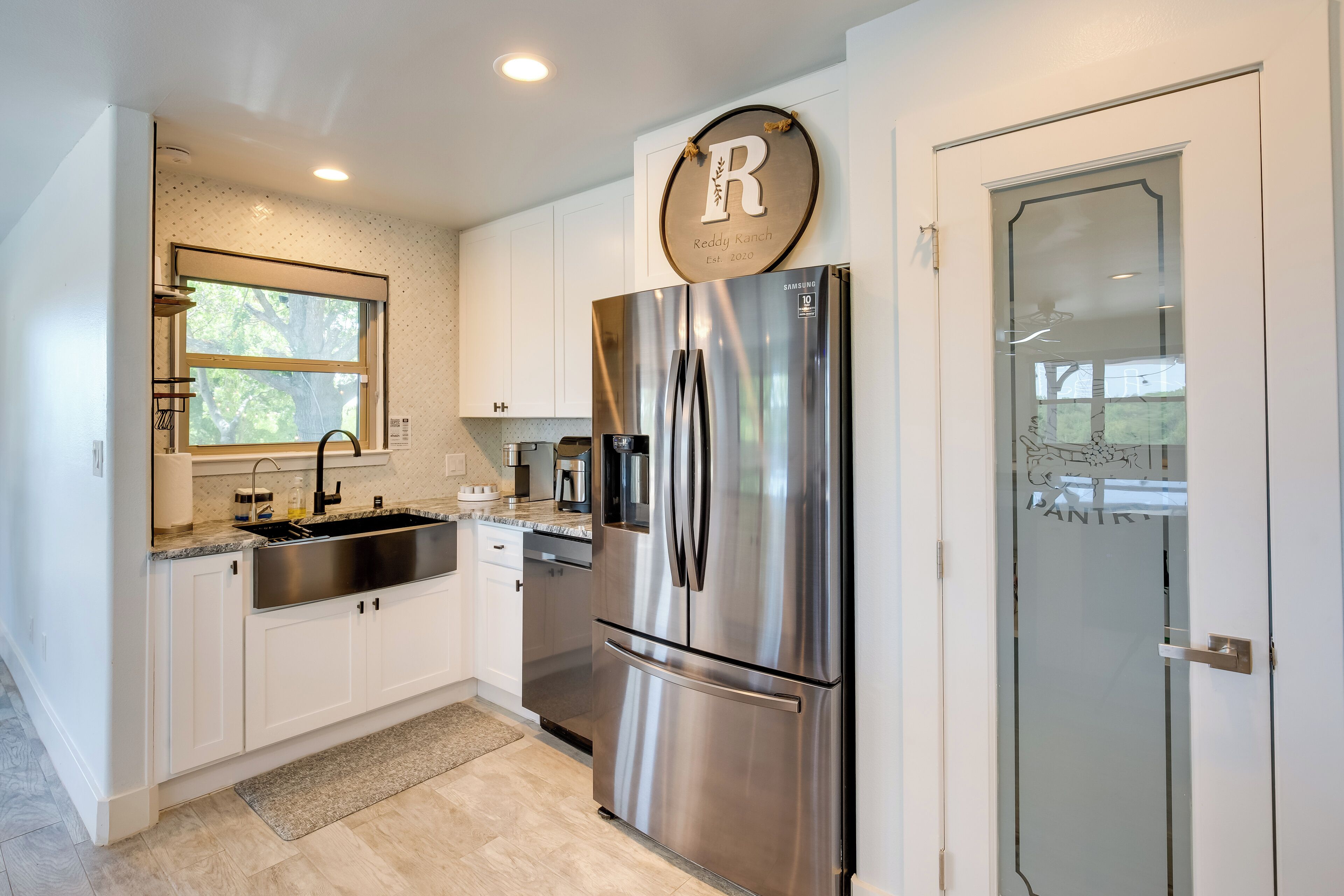 A modern kitchen with white cabinets, a stainless steel refrigerator, a window, and a door with a glass panel.