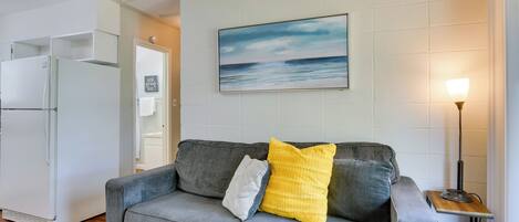 Un salon avec un canapé gris, un coussin jaune, un coussin gris à motifs, un réfrigérateur blanc, une table d’appoint en bois avec une lampe, et un tableau représentant une scène de plage accroché au mur.