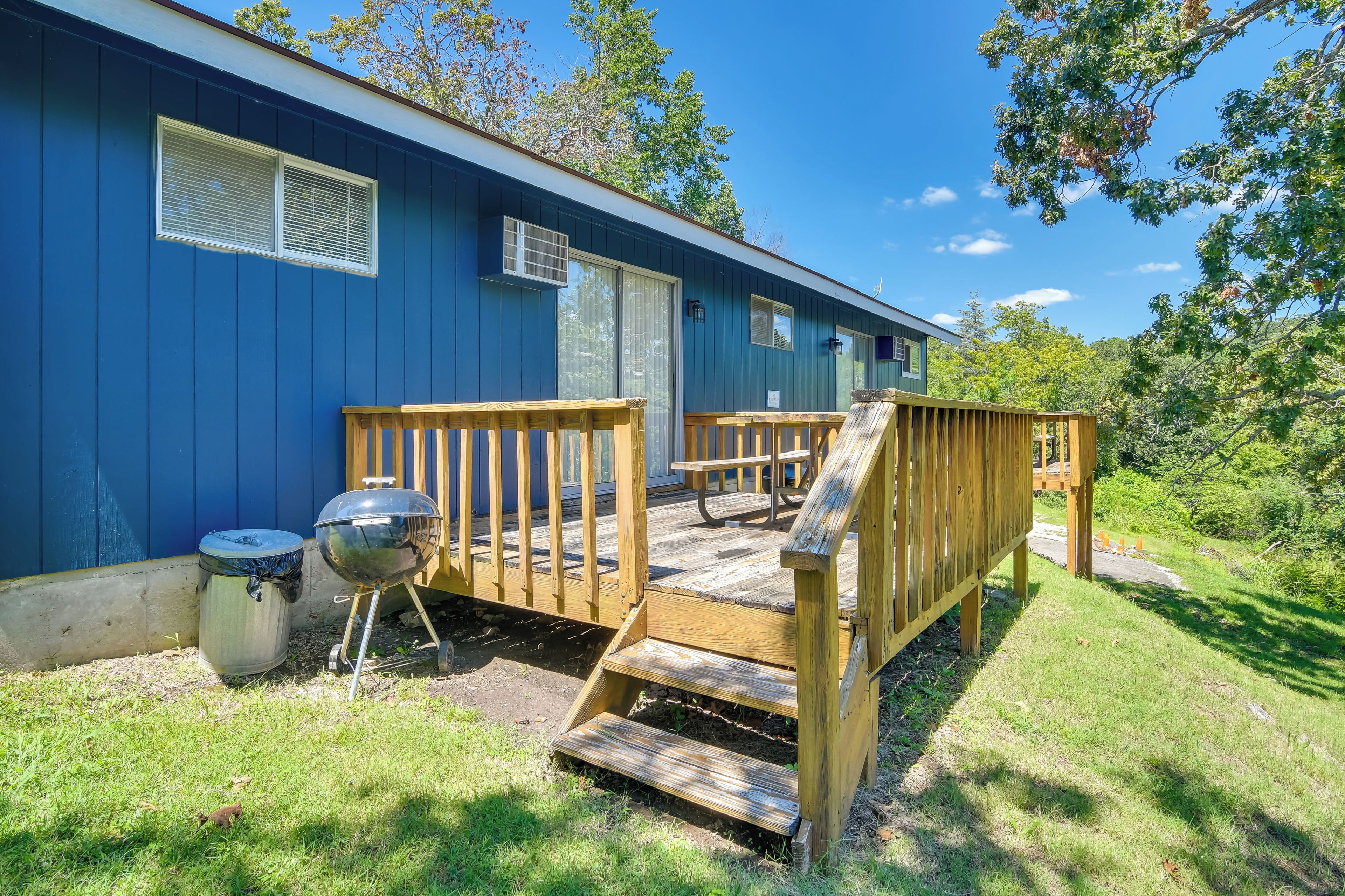 A blue mobile home with a wooden deck, a barbecue grill, and a trash can.