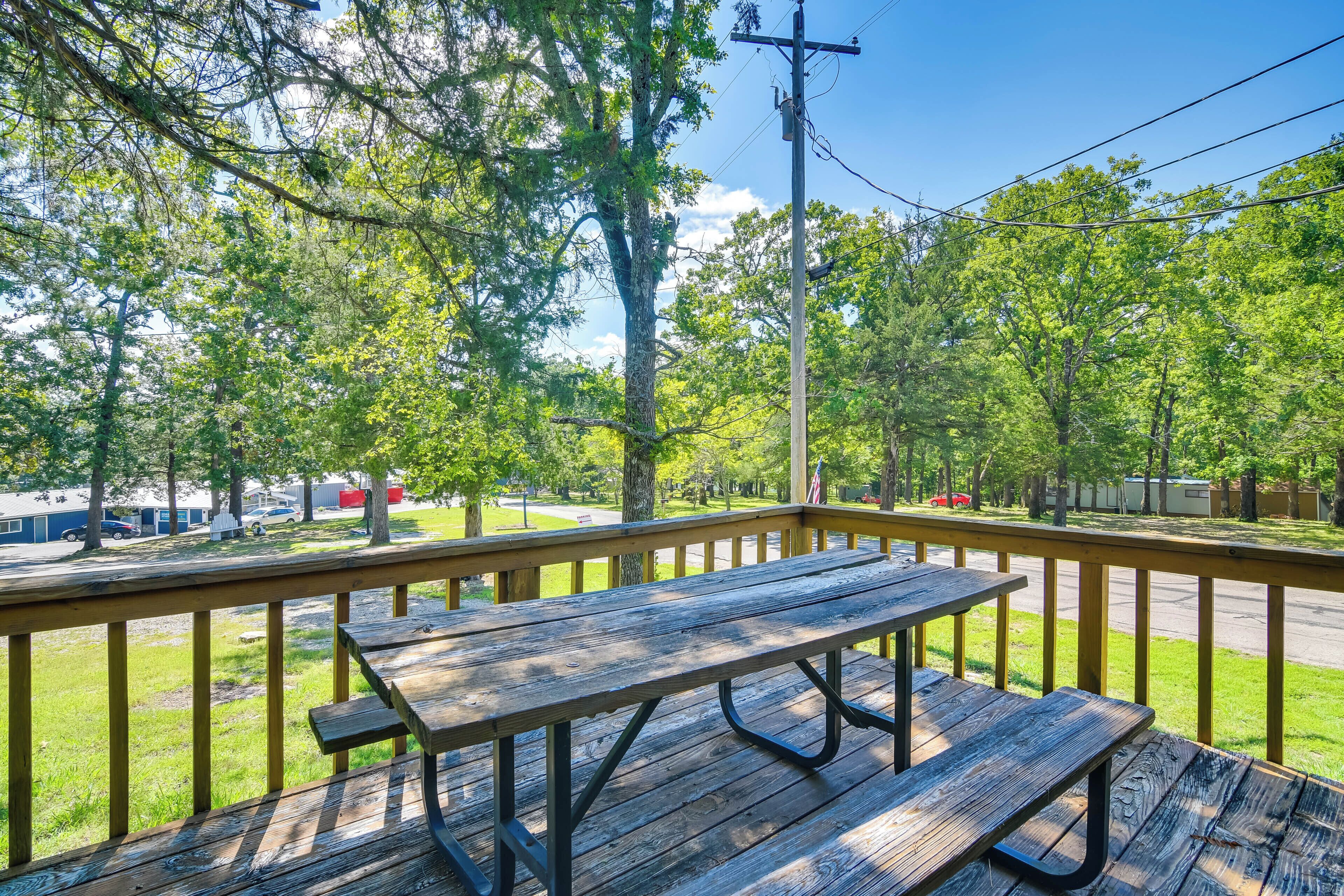 A wooden picnic table on a deck with a railing, overlooking a street with parked cars and trees.