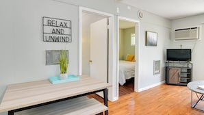 A modern living room with a wooden floor, a dining table, a TV, and a bed with a yellow pillow. - Lakefront Oasis: Boat Slip, Kayaks & Outdoor Pool (Kimberling City)