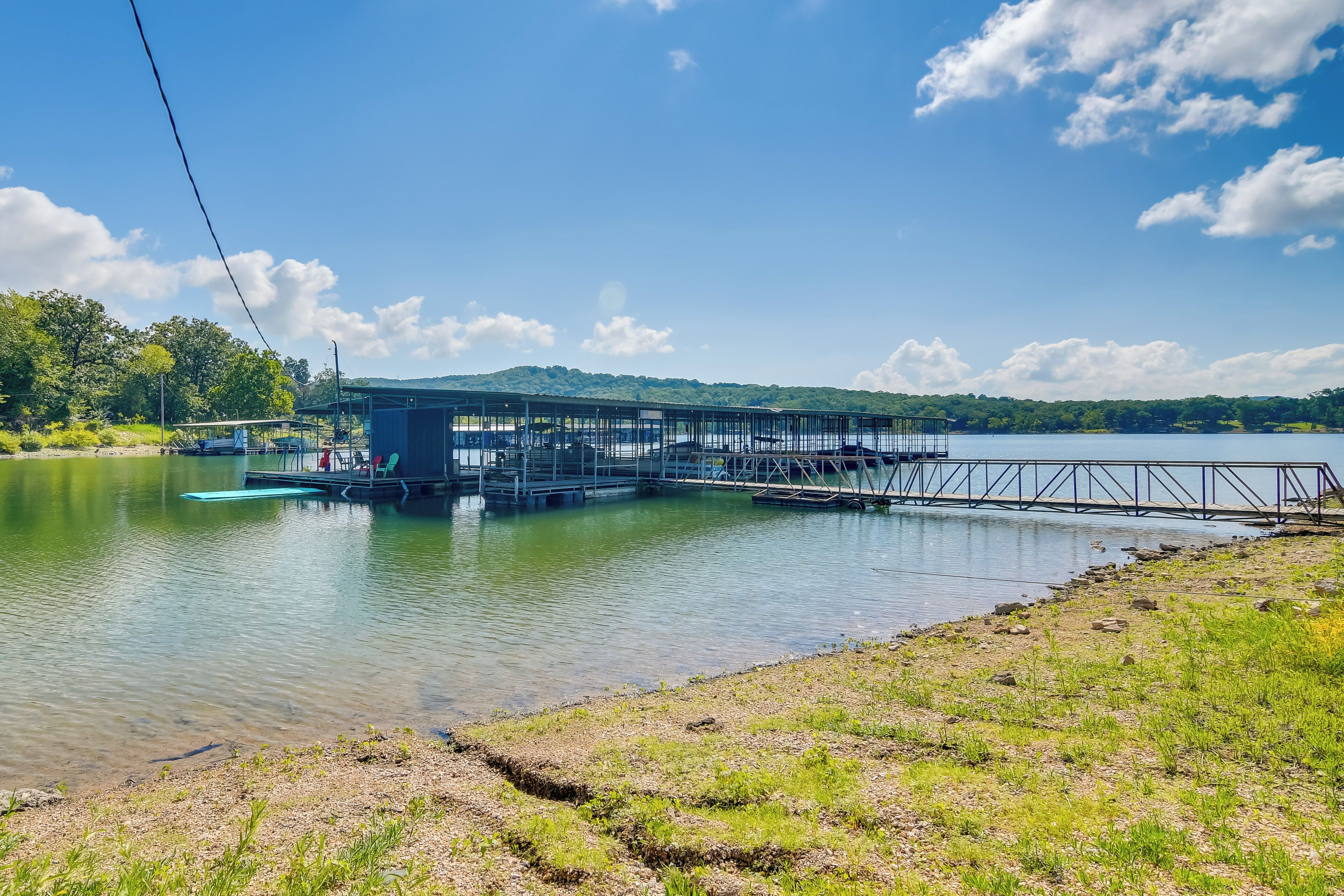 A dock extending into a calm lake with a small building on it, surrounded by greenery and hills in the background.