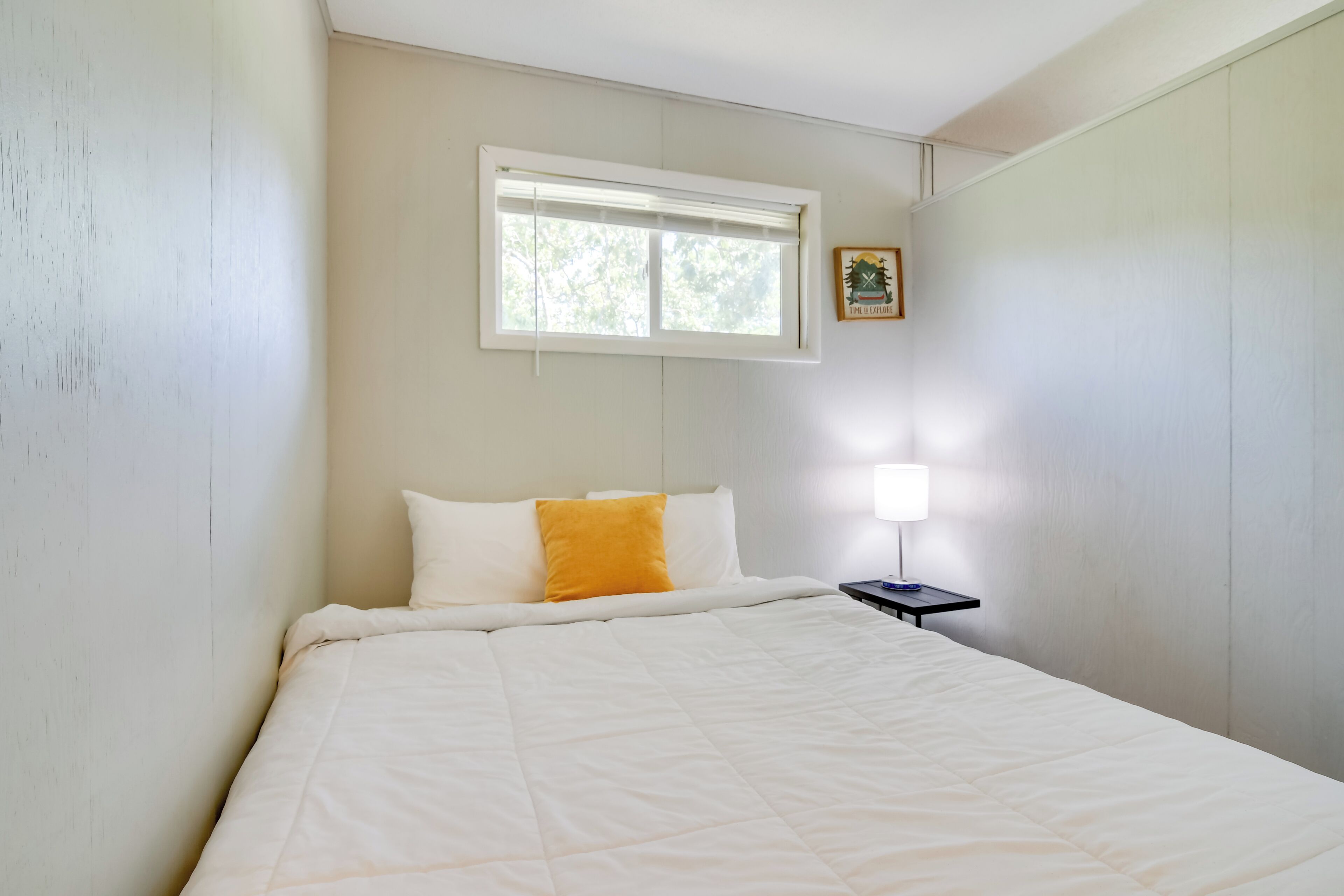 A bedroom with a bed, a window, a bedside table with a lamp, and a small picture on the wall.