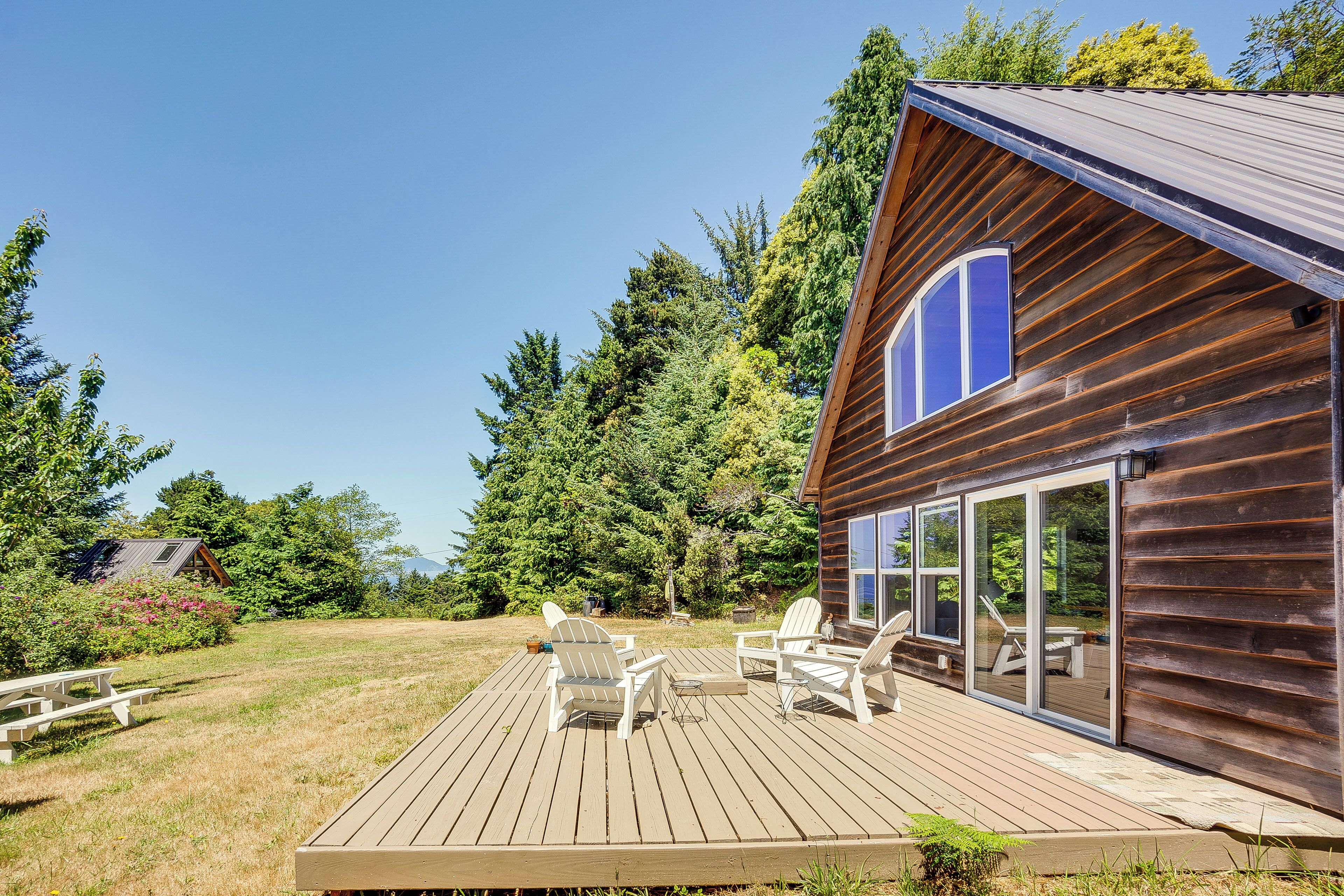 Une terrasse en bois, dotée de chaises Adirondack et d’une table de pique-nique, qui offre une vue sur une zone boisée.
