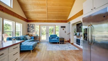 A spacious living area with a wooden ceiling, a large blue sofa, a kitchen with a stainless steel refrigerator, and a view of greenery outside.