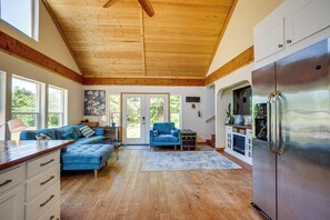 A spacious living area with a wooden ceiling, a large blue sofa, a kitchen with a stainless steel refrigerator, and a view of greenery outside.
