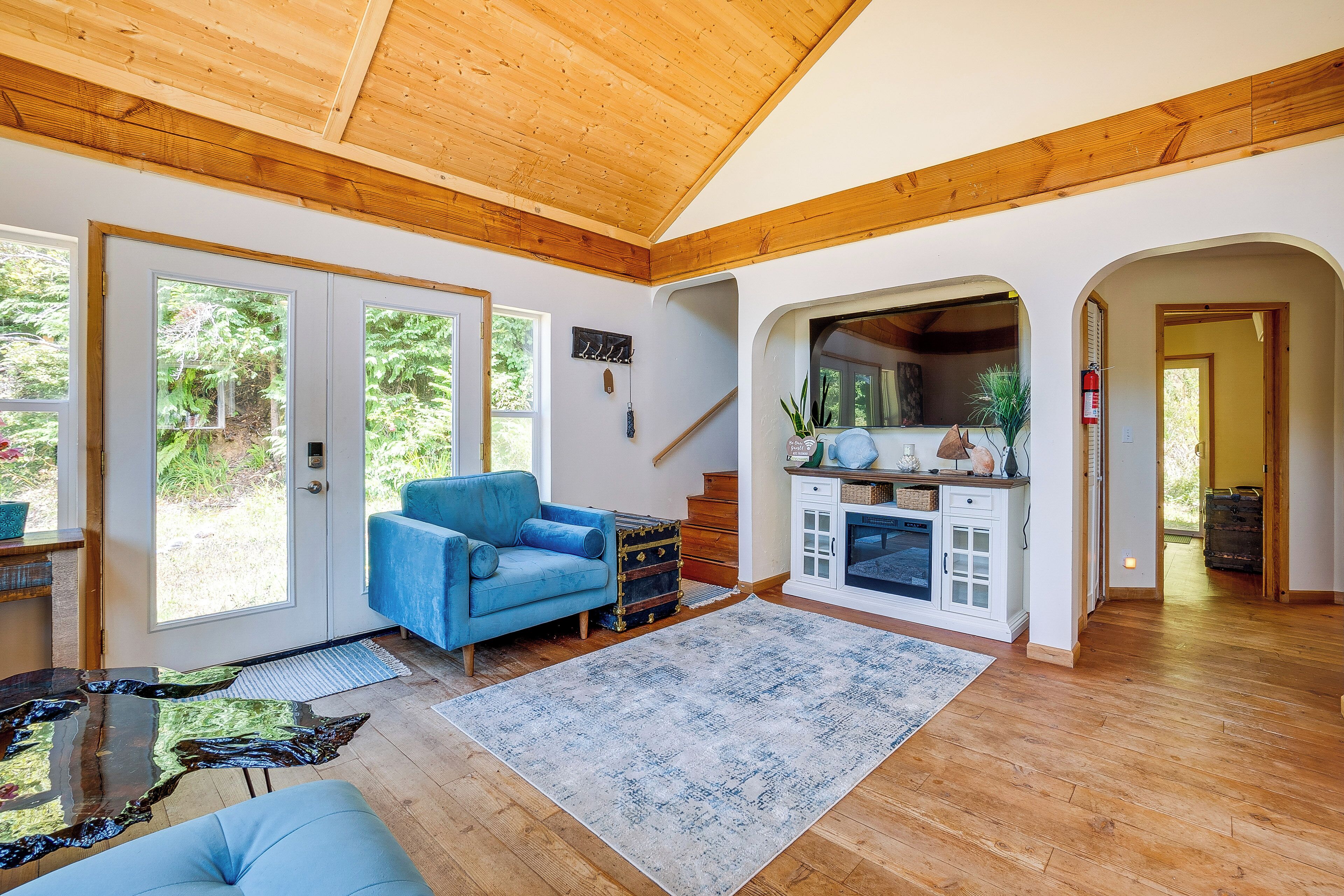 A spacious living area with a wooden ceiling, a blue sofa, a rug, and a kitchen area with cabinets and a stove.