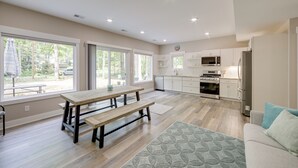 A spacious living area with a wooden table and benches, a kitchen with white cabinets and appliances, and large windows offering a view of the outdoors. - Holland Home w/ Fire Pit: Walk to Lake Michigan! (Holland)