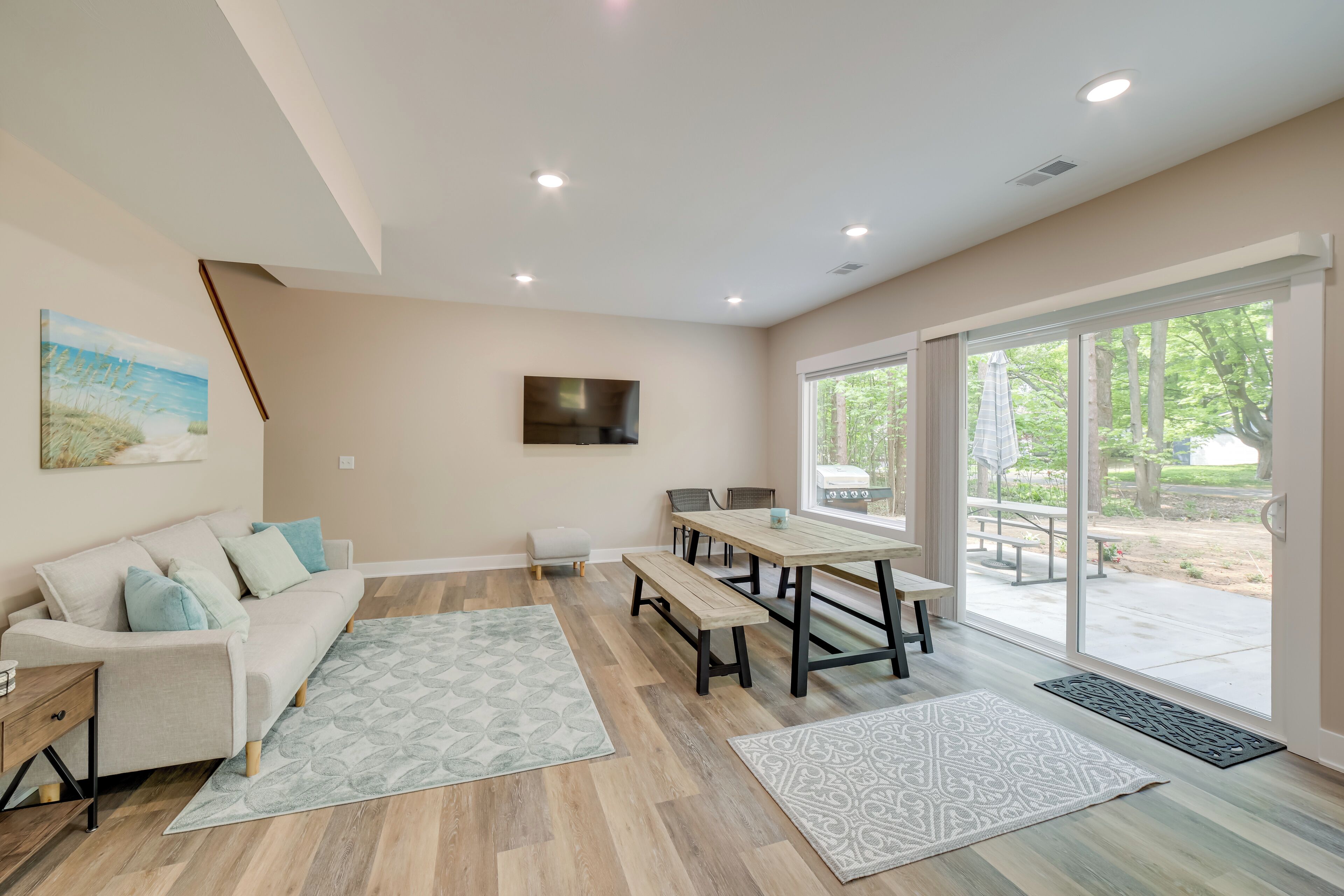 A modern living room with a sofa, coffee table, and dining table. There is a TV mounted on the wall and large windows providing a view of the outdoors.