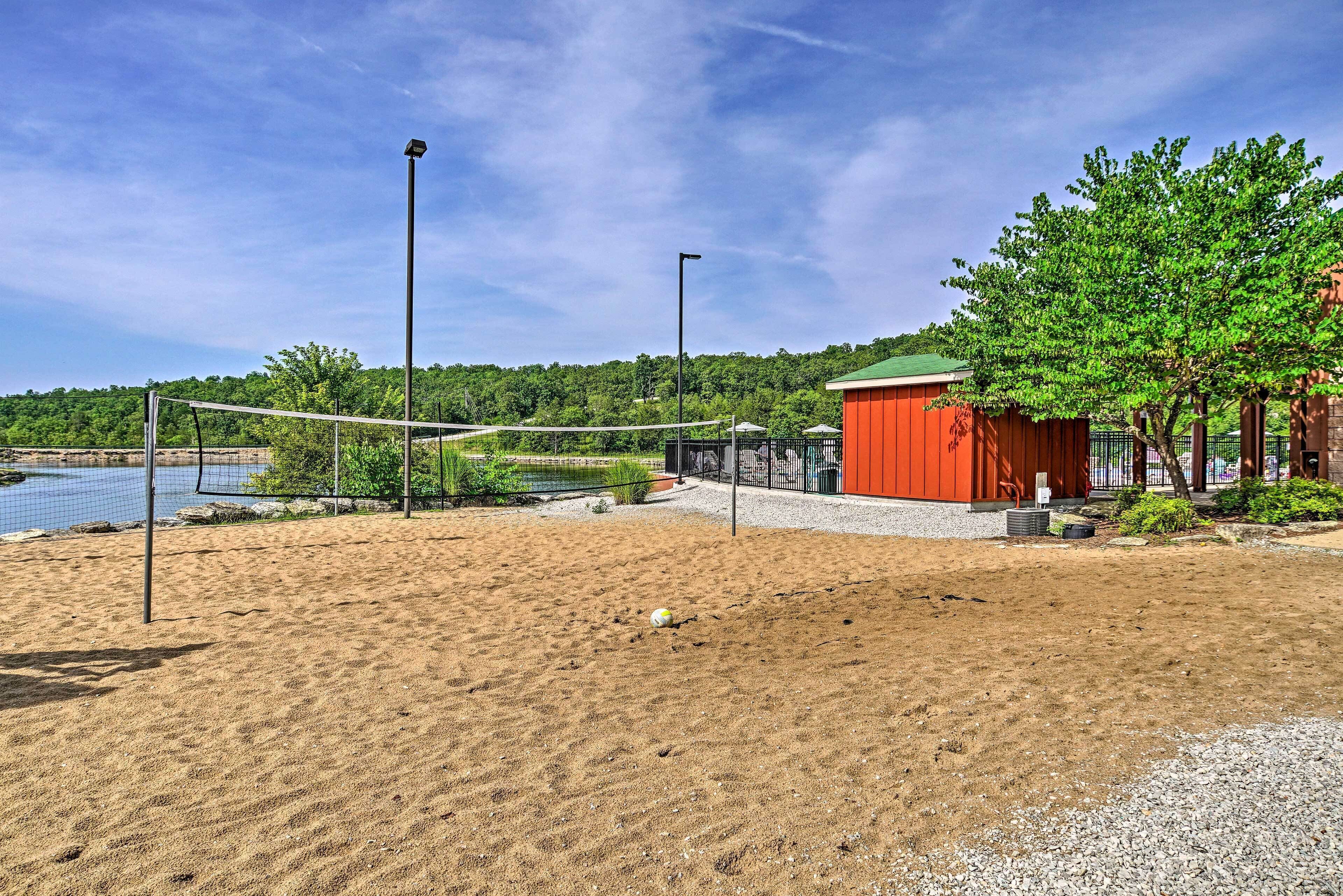 Un terrain de beach-volley sablonneux avec un filet, un bâtiment rouge et un arbre en arrière-plan.