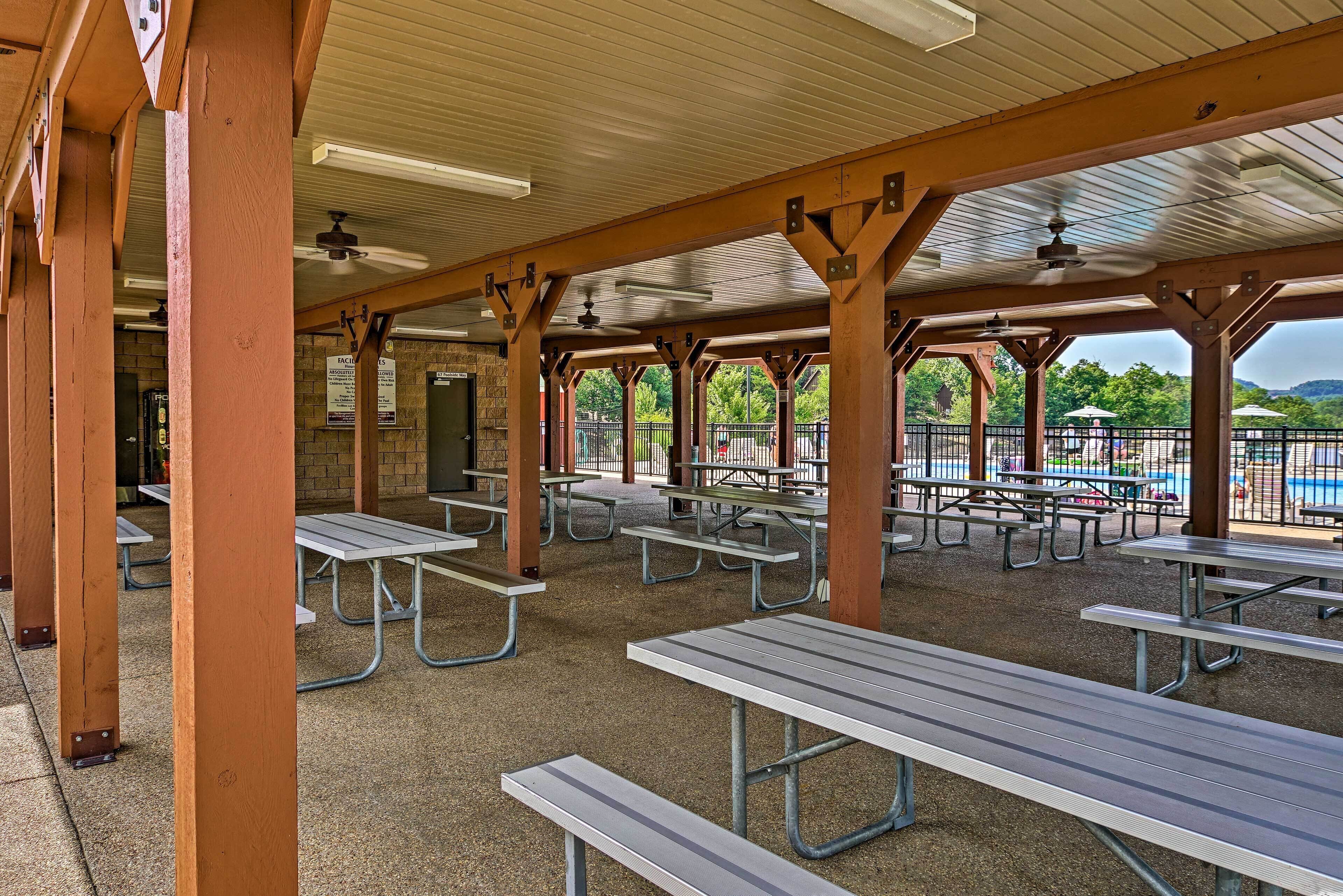 A covered picnic area with metal tables and benches, a brick wall, and a view of a playground and trees.