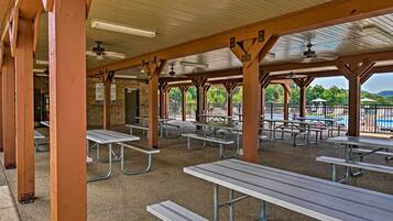 A covered picnic area with metal tables and benches, a brick wall, and a view of a playground and trees.