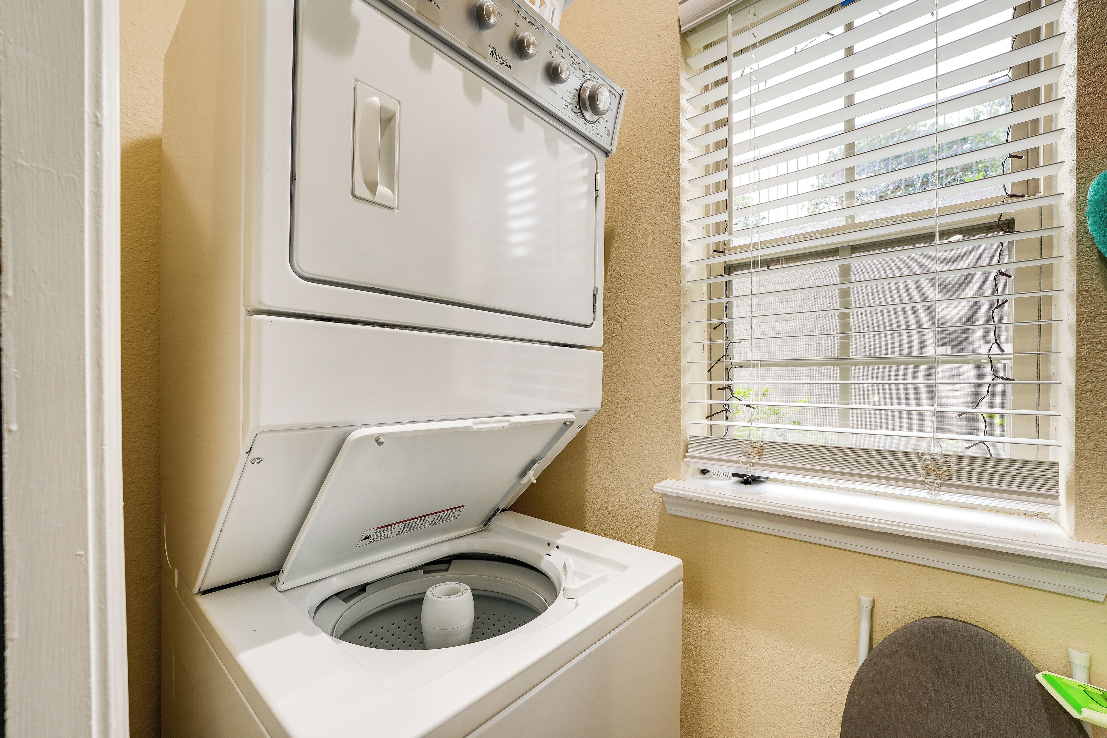 A laundry area with a stacked washer and dryer, a window with blinds, and a green sign on the wall.