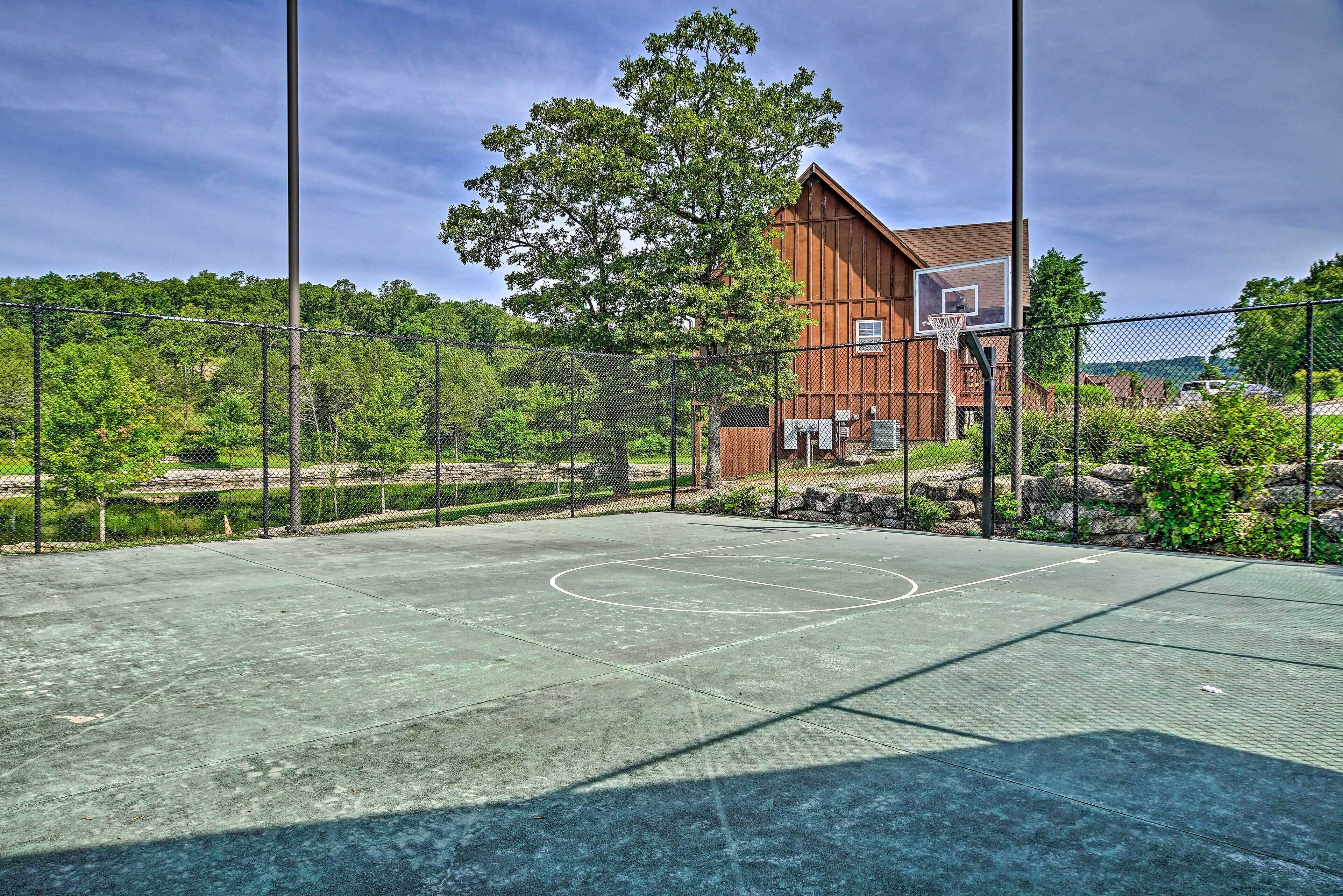 An outdoor basketball court with a fence, trees, and a building in the background.