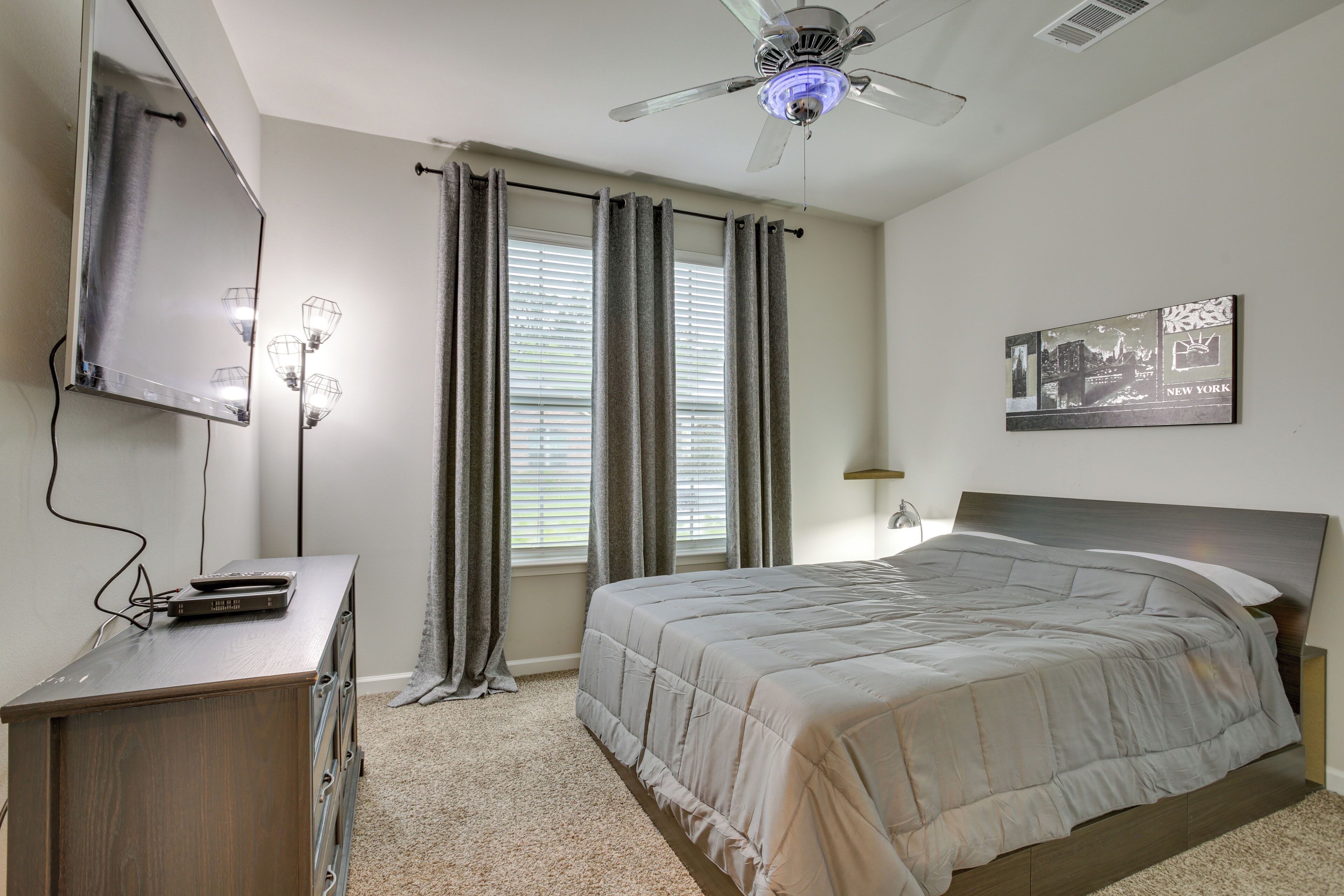 Quarto com cama, ventoinha no teto, televisão presa na parede e janela com cortinas.