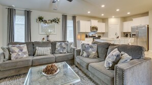 A modern living room with a glass coffee table, two grey sofas with patterned cushions, a ceiling fan, and a kitchen in the background. - Baton Rouge Vacation Rental: 5 Mi to Lsu! (Baton Rouge)