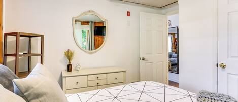 Une chambre à coucher avec une couvre-lit à motifs géométriques, une commode, un miroir et une porte donnant sur une autre pièce.