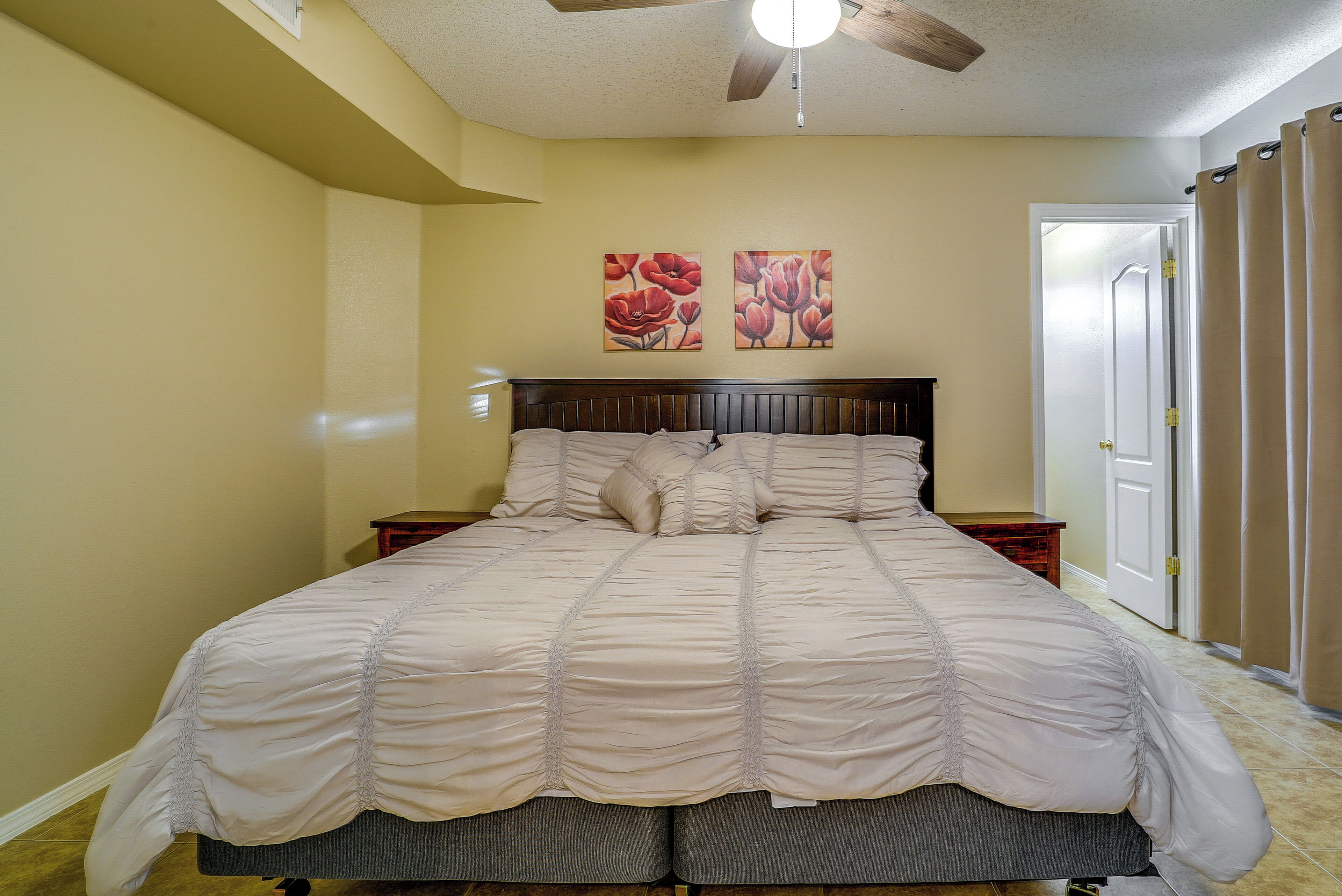 A bedroom with a large bed, two nightstands, a ceiling fan, and two floral paintings on the wall.