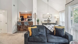 Una sala de estar moderna con un sofá oscuro, una zona de comedor con mesa y sillas, y una cocina al fondo.