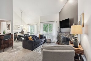 A modern living room with a dark sofa, a grey armchair, a dining area with a table and chairs, a fireplace, and a TV mounted on the wall. - Overland Park Condo, Close to Lakes & Parks! (Overland Park)