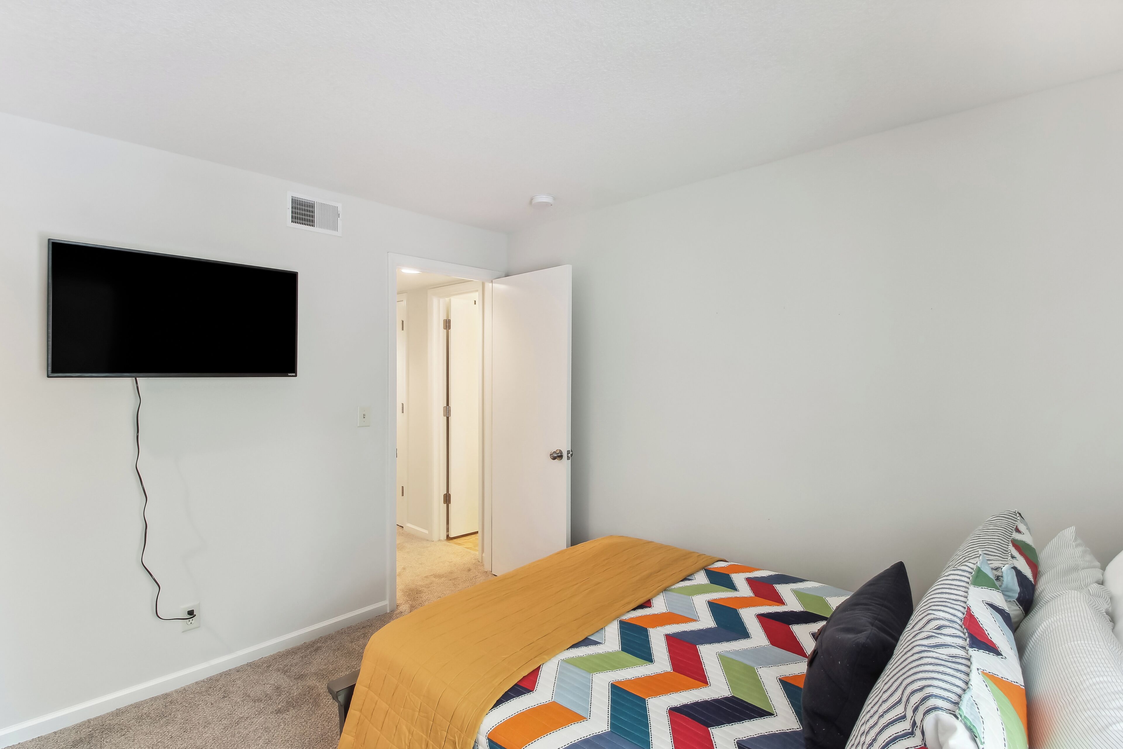 Un dormitorio con cama, televisor montado en la pared y una puerta que da a otra habitación.
