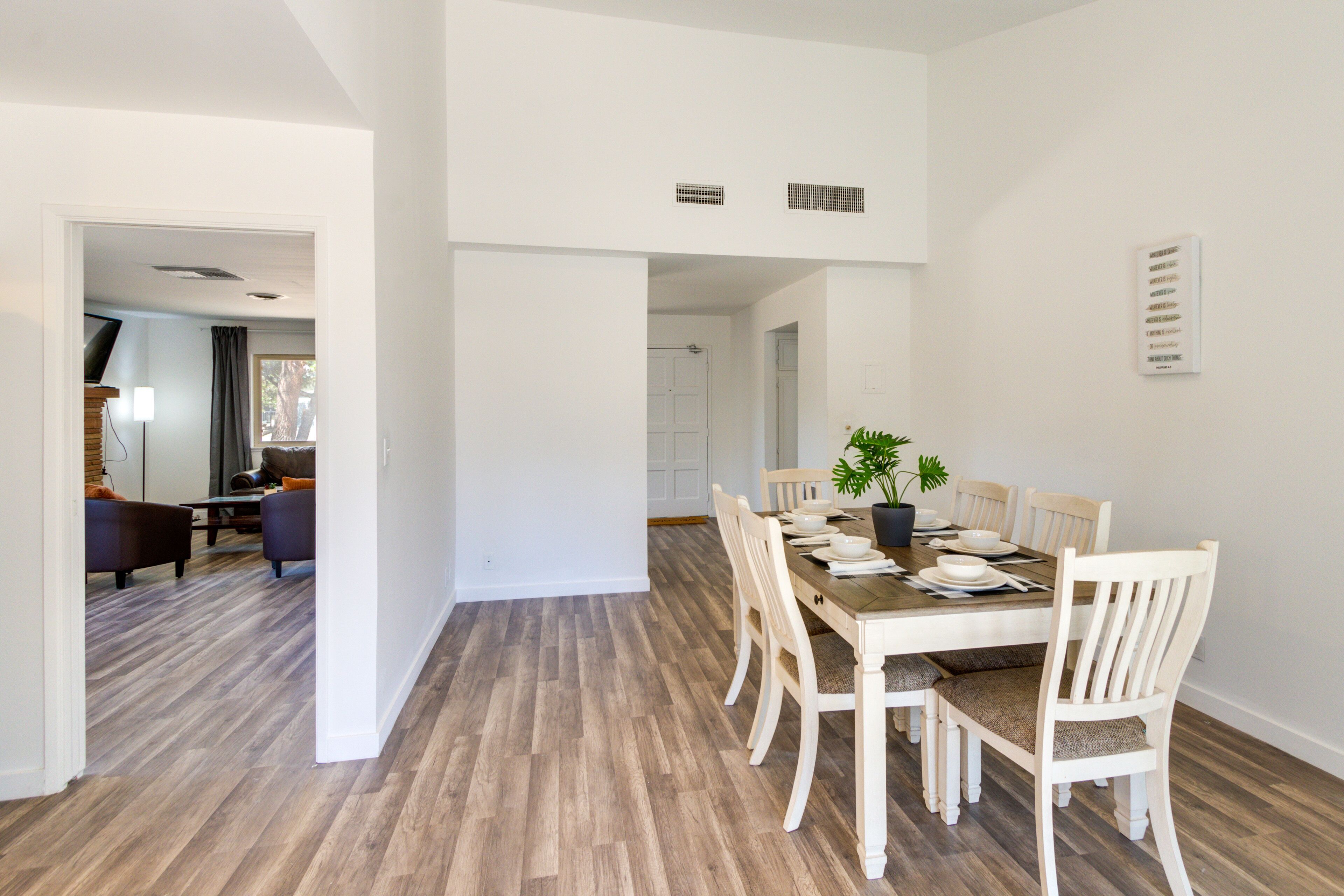 Un comedor con mesa de madera y sillas blancas, una planta en maceta y una pared blanca con un respiradero.