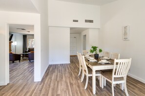 Un comedor con mesa de madera y sillas blancas, una planta en maceta y una pared blanca con un respiradero.