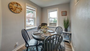 A dining room with a round table, black chairs, a wall clock, and two windows with blinds.