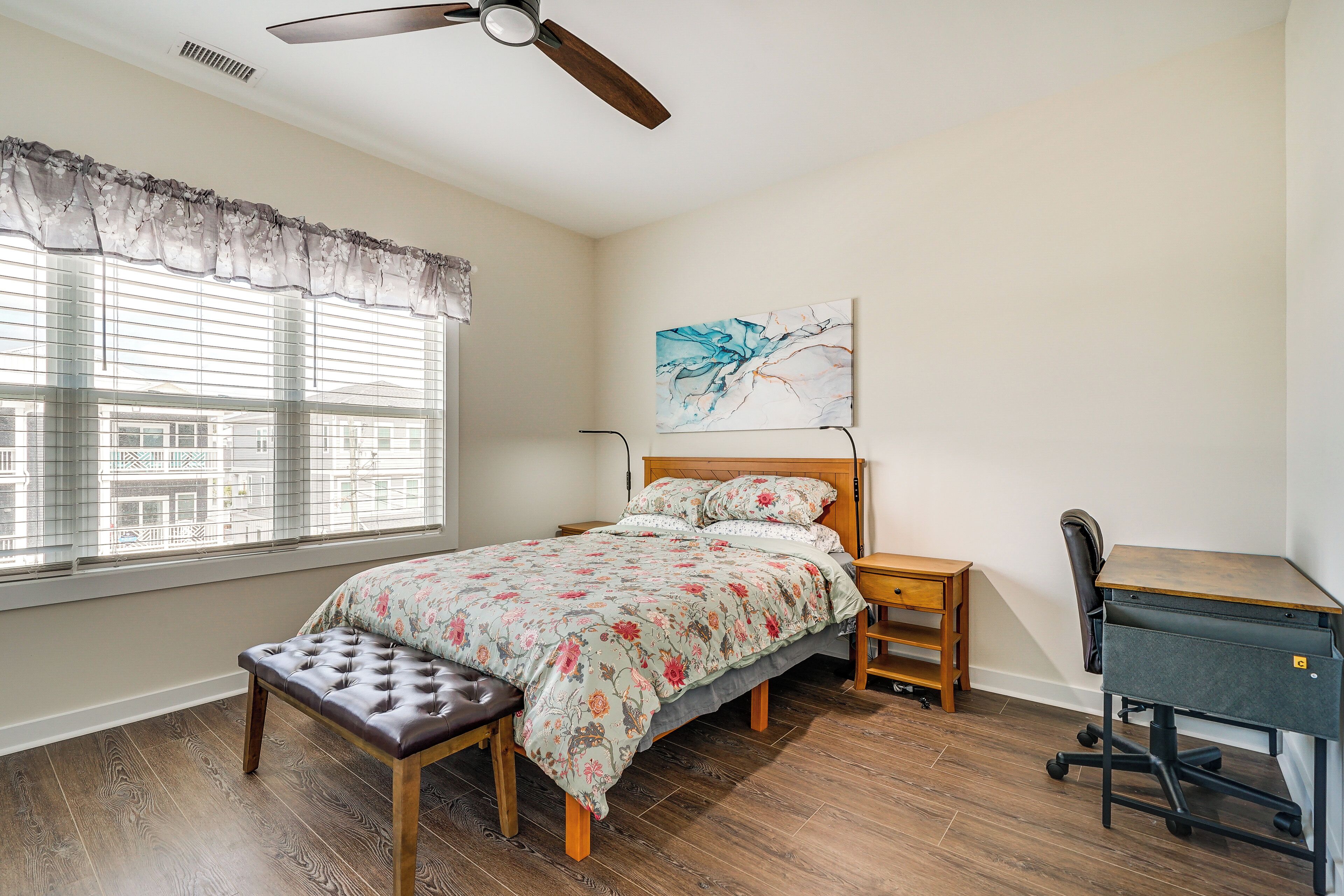 Une chambre à coucher comprenant un lit, un bureau, une chaise, un banc, un ventilateur de plafond et une fenêtre avec des stores.