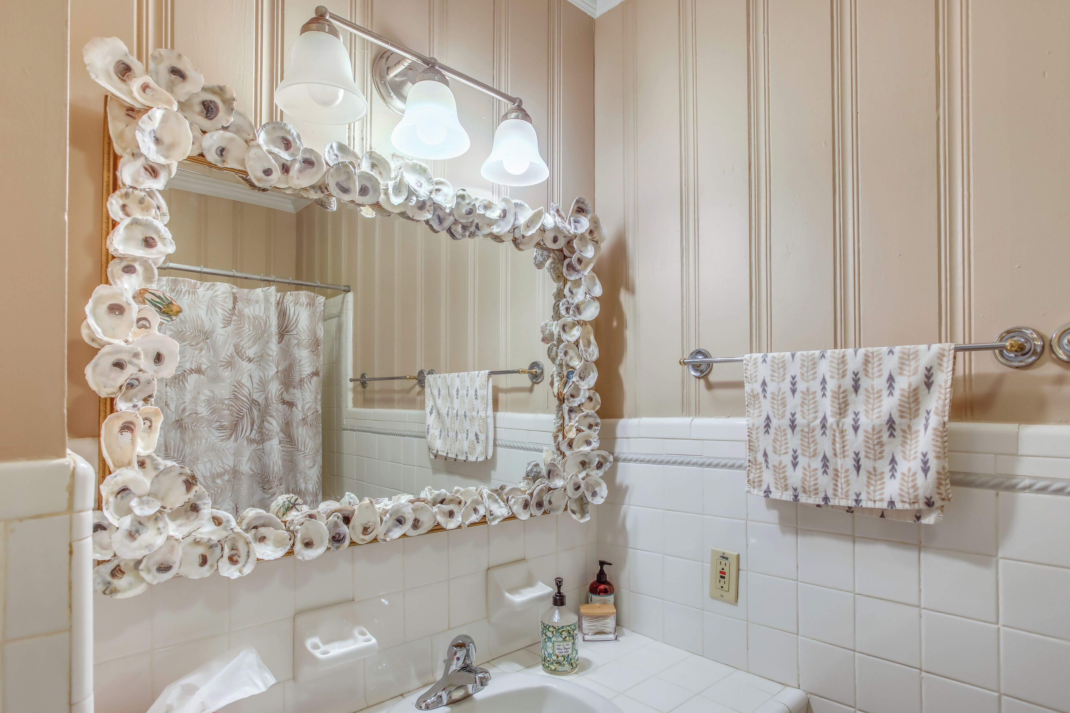 Une salle de bain avec un miroir encadré de coquilles d’huîtres, un lavabo blanc et un porte-serviettes avec deux serviettes.