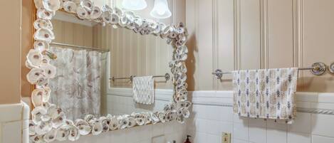 Une salle de bain avec un miroir encadré de coquilles d’huîtres, un lavabo blanc et un porte-serviettes avec deux serviettes.