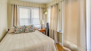 A bedroom with a bed, curtains, a window with blinds, a bedside table with a lamp, and a hanging plant. - Carolina Beach Home w/ Private Deck: Walk to Shore (Carolina Beach)