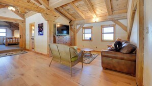 Un salon spacieux avec des poutres en bois, un canapé en cuir marron, un fauteuil en osier, une petite table ronde et un téléviseur.