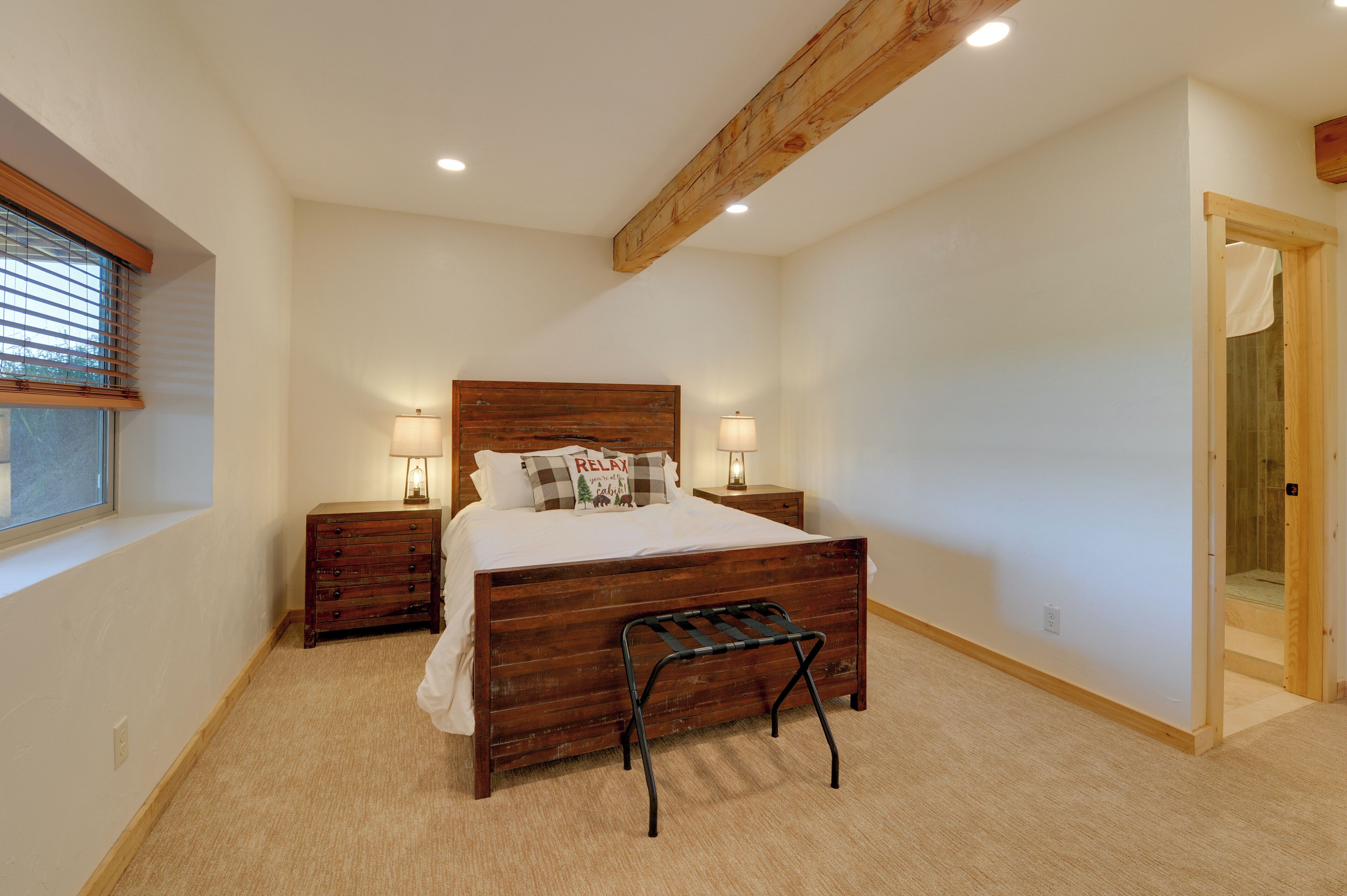 A bedroom with a wooden bed, two bedside tables with lamps, a bench at the foot of the bed, and a window with blinds.