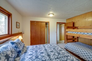 A bedroom with a bunk bed, a bed with patterned bedding, a wooden wardrobe, and a window with a view of trees.