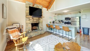 A modern kitchen with a stone fireplace, wooden ceiling beams, and a bar area with bar stools. - Wooded Retreat w/ Decks & Views in Beech Mountain! (Banner Elk)