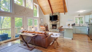 A spacious living area with a wooden ceiling, a stone fireplace, a large sofa, and a kitchen with light blue cabinets. - Wooded Retreat w/ Decks & Views in Beech Mountain! (Banner Elk)