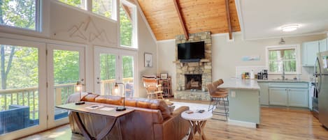 A spacious living area with a wooden ceiling, a stone fireplace, a large sofa, and a kitchen with light blue cabinets.