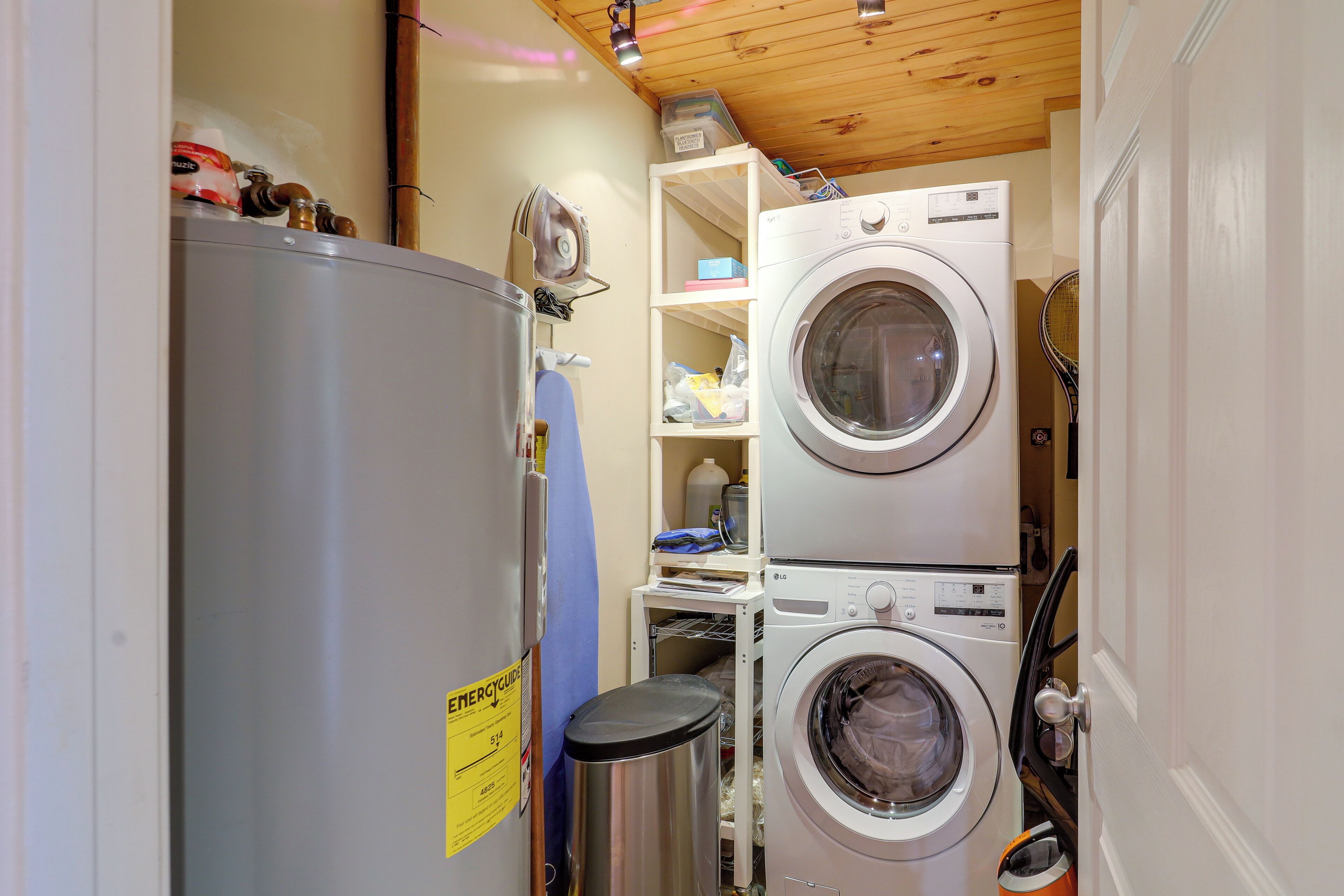 Une petite buanderie comprenant une machine à laver et un sèche-linge superposés, une poubelle en acier inoxydable et une petite étagère contenant divers objets.