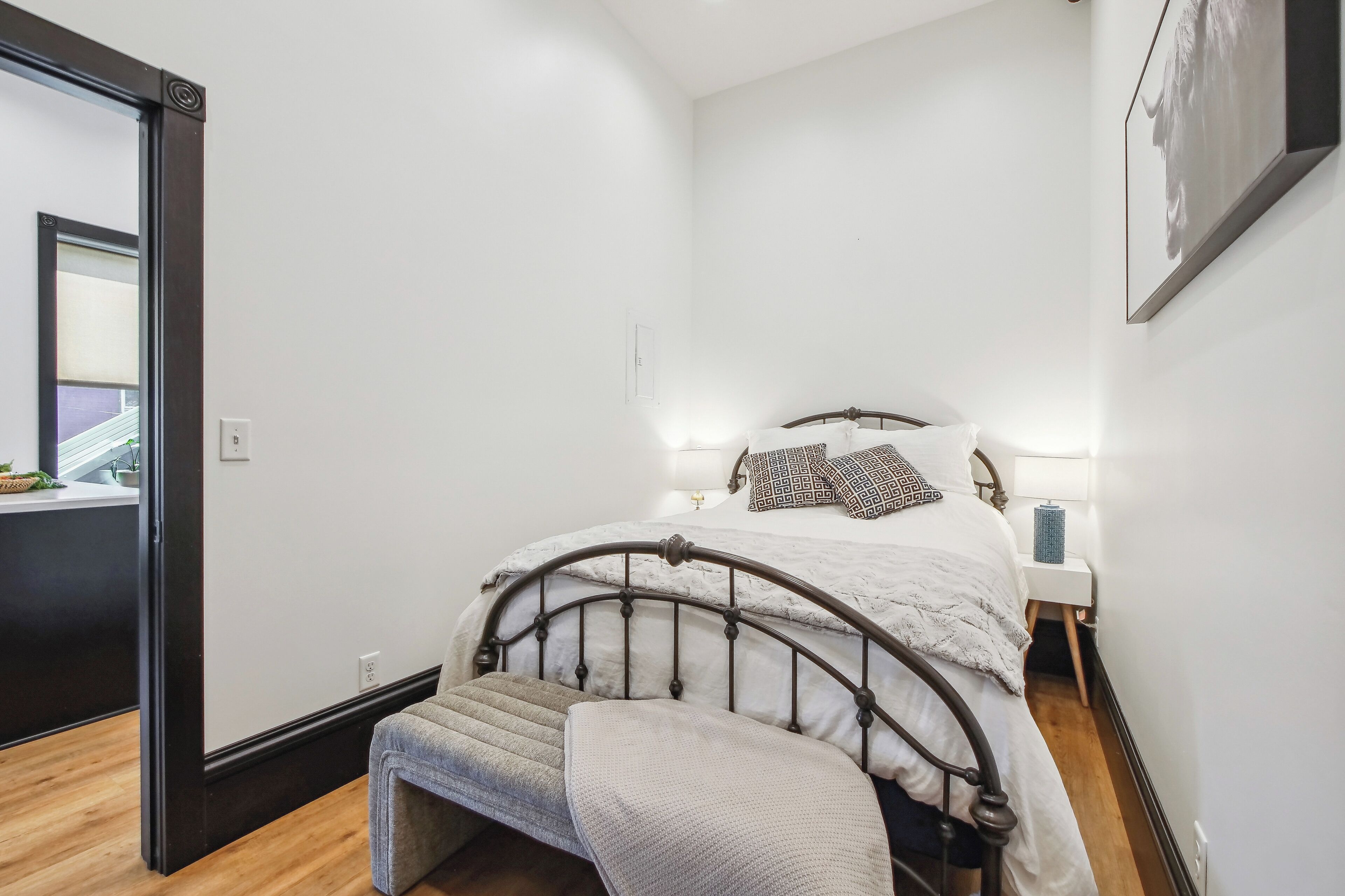 A bedroom with a metal bed frame, white bedding, a bedside table with a lamp, and a wall-mounted picture.