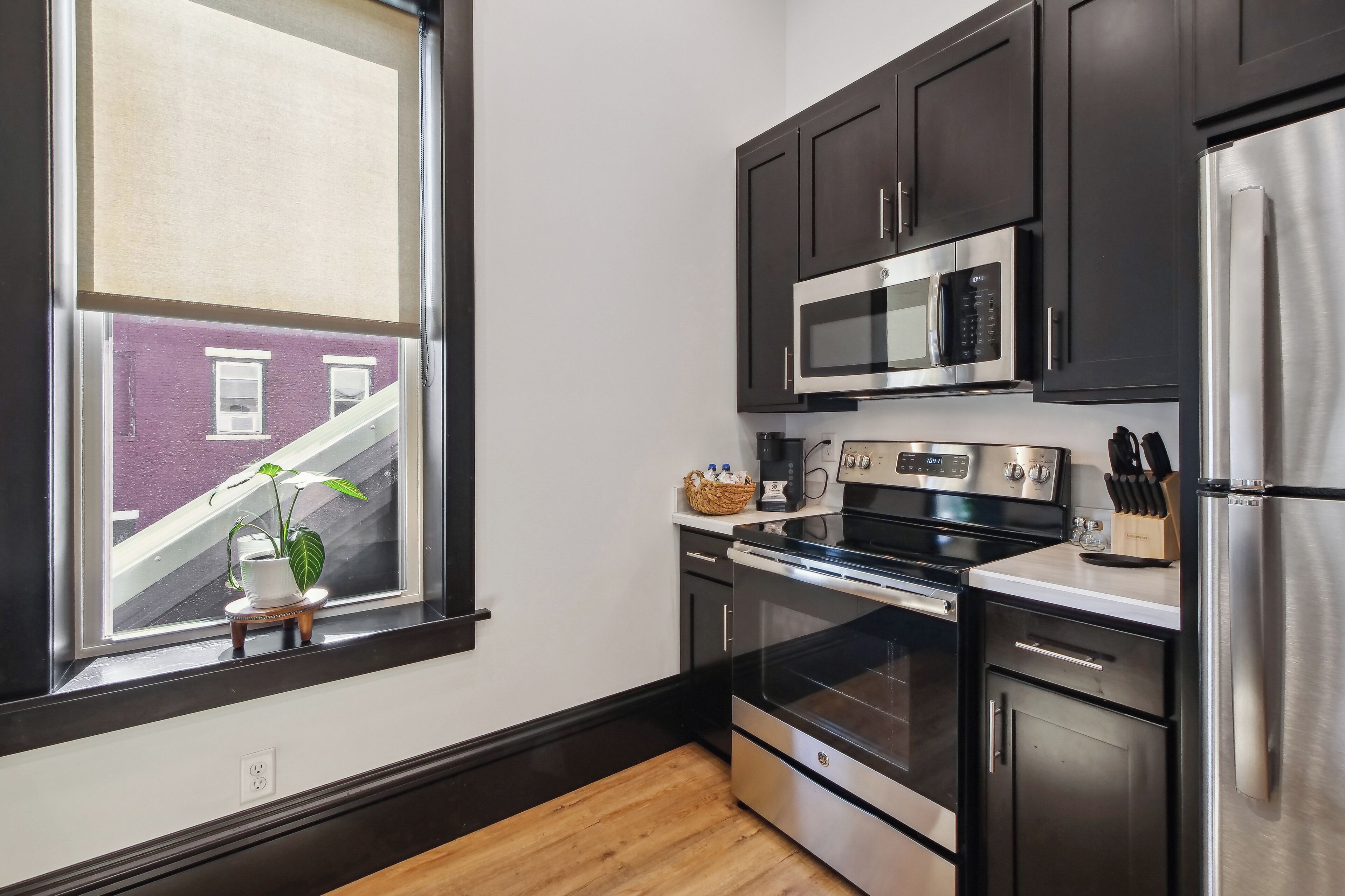 A modern kitchen with dark cabinets, stainless steel appliances, and a window with a view of a building outside.