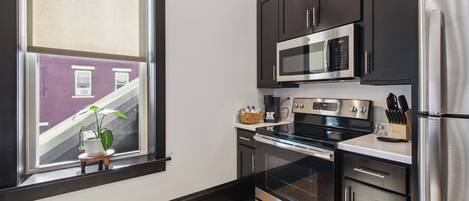 A modern kitchen with dark cabinets, stainless steel appliances, and a window with a view of a building outside.