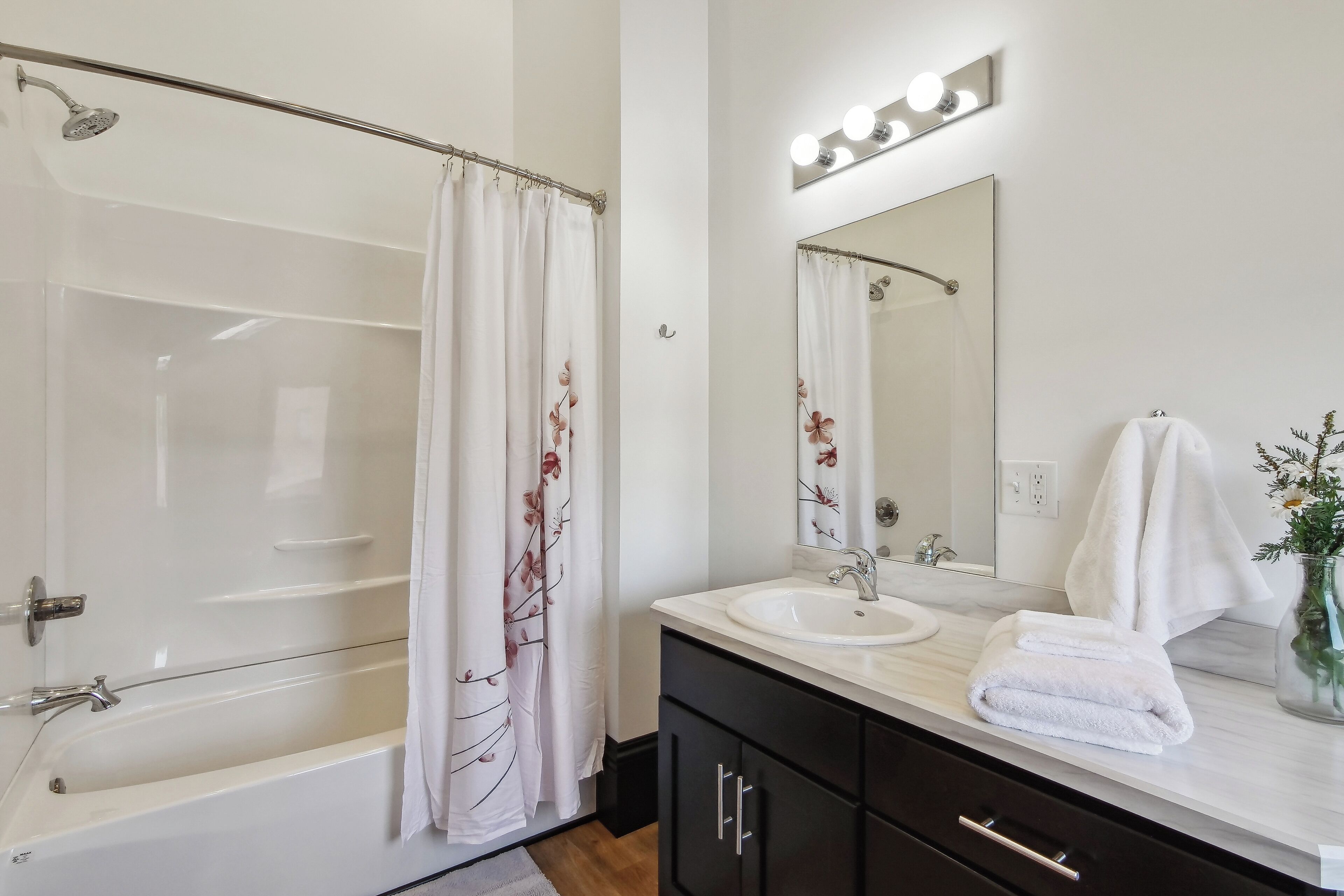 A modern bathroom with a bathtub, a sink, a mirror with lights, and a shower curtain with floral patterns.